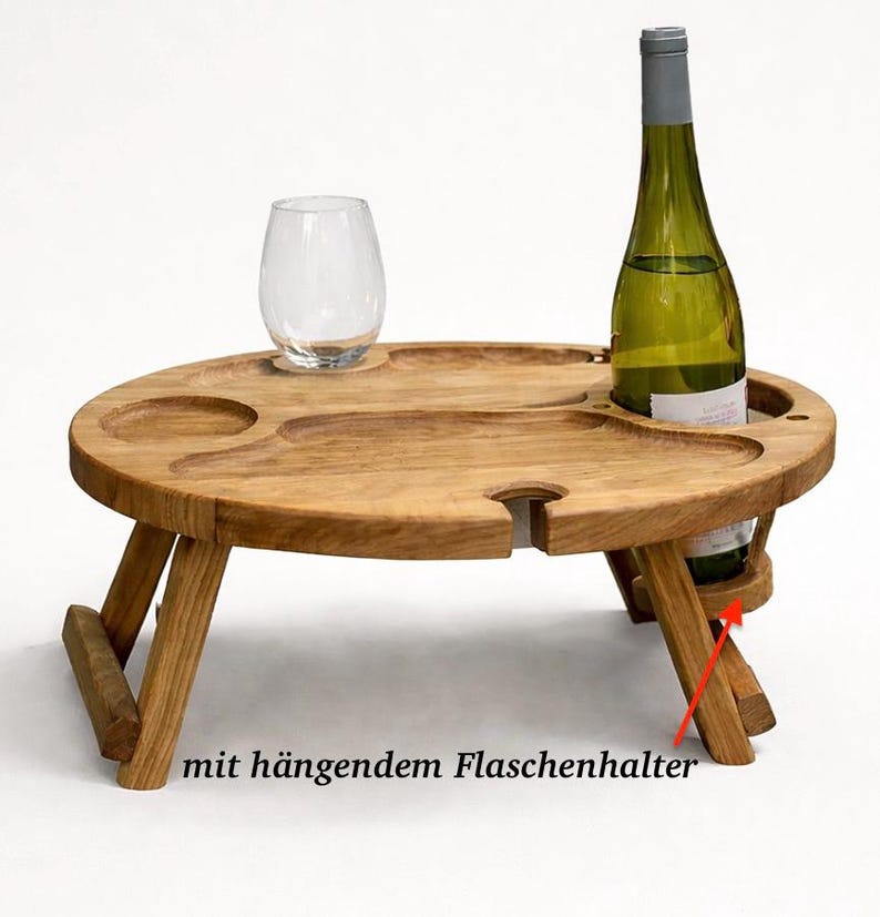 Op de afbeelding: Een ronde inklapbare houten tafel met uitgesneden inkepingen voor een glas en een fles. De tafel heeft een hangende flessenhouder aan de zijkant, aangegeven door een rode pijl en de Duitse tekst "mit h&auml;ngendem Flaschenhalter".