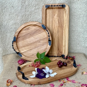 Può includere: Tre vassoi di legno di diverse forme: un vassoio rotondo, un vassoio rettangolare e un vassoio ovale. Ogni vassoio ha manici e dettagli decorativi in metallo nero. I vassoi sono esposti con fiori e bacche.