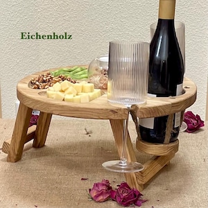 Pode incluir: Uma mesa de piquenique de madeira com tampo redondo e pernas dobráveis. A mesa tem recortes para uma garrafa de vinho e uma taça de vinho. Contém queijo, nozes e kiwi. O texto "Eichenholz" é visível no canto superior esquerdo.