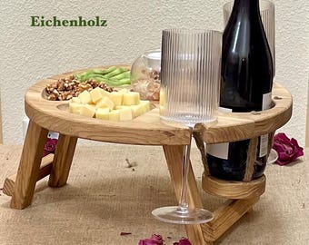 May include: A wooden picnic table with a round top and folding legs. The table has cutouts for a wine bottle and a wine glass. It holds cheese, nuts, and kiwi. The text "Eichenholz" is visible on the top left.