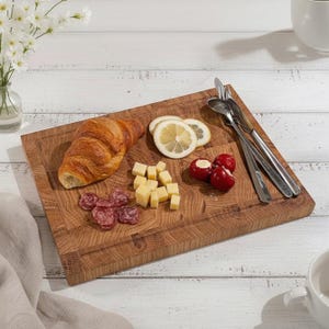 May include: A wooden serving board with a croissant, cheese cubes, salami, lemon slices, and stuffed cherry peppers. Silverware rests on the board. The board is made of light brown wood and is on a white wooden surface.