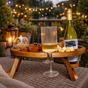 Op de afbeelding: Een ronde inklapbare houten snacktafel met een centrale uitsparing voor de steel van een wijnglas. De tafel bevat een fles witte wijn, een glas witte wijn, een glazen kaarsenhouder met een brandend waxinelichtje en gedroogd fruit en kaas.
