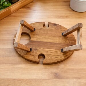 Op de afbeelding: Een ronde houten wijntafel met een natuurlijke houtnerf, voorzien van vier inklapbare poten en twee ronde uitsparingen op het oppervlak voor wijnglazen en een fles, weergegeven in omgekeerde positie met de poten naar boven geklapt.