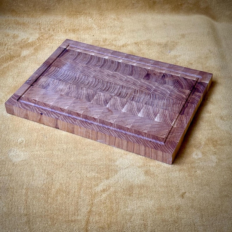 May include: A rectangular end-grain butcher block cutting board made of light brown wood. The board has a juice groove around the perimeter. The wood grain pattern is visible throughout the board, creating a textured surface. This kitchen item is ideal for food preparation.