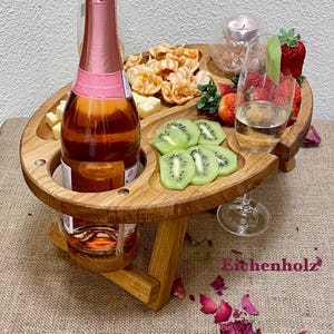 Könnte beinhalten: Ein hölzerner Picknicktisch mit integrierten Flaschenhaltern und einem Servierbereich. Der Tisch ist mit einer Flasche Rosé-Champagner, zwei Gläsern, Kiwischeiben, Erdbeeren und Crackern gedeckt. Der Tisch ist aus Eichenholz gefertigt und hat eine natürliche Oberfläche.