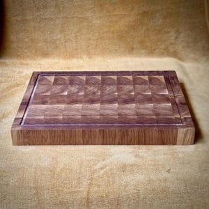 May include: A rectangular end-grain butcher block cutting board. The board is made of light and dark brown wood blocks arranged in a checkerboard pattern. A juice groove runs around the perimeter of the cutting surface. The board is approximately 16 inches long and 12 inches wide.