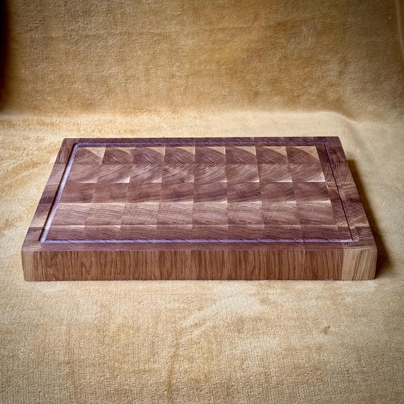 May include: A rectangular end-grain butcher block cutting board. The board is made of light and dark brown wood blocks arranged in a checkerboard pattern. A juice groove runs around the perimeter of the cutting surface. The board is approximately 16 inches long and 12 inches wide.