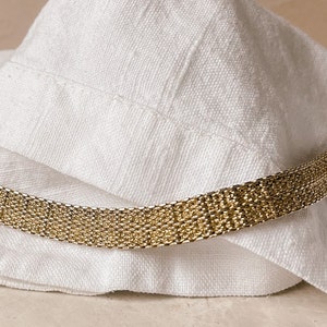 Puede incluir: Un brazalete de cadena dorado con un diseño delicado y tejido. El brazalete está sobre una superficie de tela blanca.