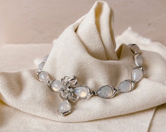 May include: A silver necklace with a teardrop-shaped pendant and a flower-shaped charm. The necklace is made of white stones and is on a white fabric background.