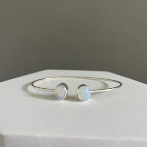 May include: A silver open cuff bracelet featuring two round, pale blue-white opal stones. The bracelet is presented on a white surface against a neutral grey background. The opal stones have a subtle, iridescent sheen.