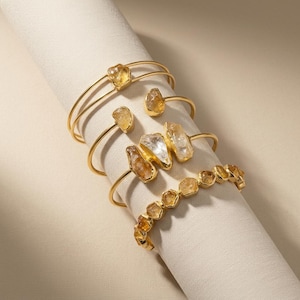 May include: A collection of four gold-toned metal cuff bracelets featuring raw, uncut yellow and clear crystals. The bracelets vary in design, with some showcasing single stones, others featuring clusters of three crystals, and one with a continuous row.