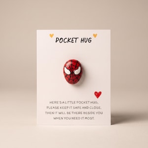 Könnte beinhalten: Eine weiße Karte mit dem Text POCKET HUG, zwei kleinen gelben Herzen und einem roten Herz. Ein kleiner, runder, roter Stein, der mit einem schwarzen Spinnennetz-Muster und weißen Augen bemalt ist, ist in der Mitte befestigt. Die Karte enthält den gedruckten Text: HERE'S A LITTLE POCKET HUG, PLEASE KEEP IT SAFE AND CLOSE, THEN IT WILL BE THERE BESIDE YOU WHEN YOU NEED IT MOST.