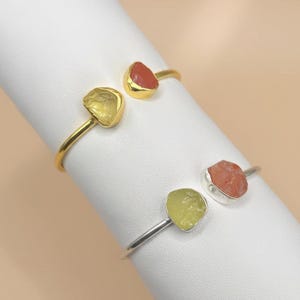 Könnte beinhalten: Zwei offene Armreifen mit rohen Edelstein-Akzenten. Ein Armreif hat eine goldene Oberfläche mit einem gelben und einem roten Edelstein. Der andere Armreif hat eine silberne Oberfläche mit einem gelben und einem roten Edelstein.