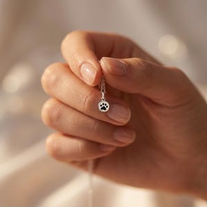 May include: A silver necklace featuring a small, circular pendant with a black paw print design. The pendant is attached to a delicate silver chain, showcasing a simple and clean aesthetic with a polished metallic finish on the circular charm.