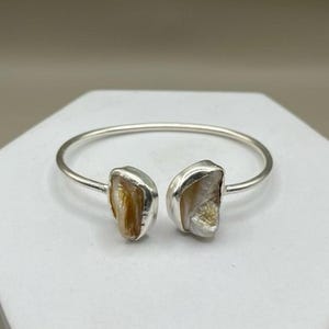Può includere: Un bracciale aperto in argento con due pietre irregolari dall'aspetto madreperlaceo e dalla consistenza grezza alle estremità. Il cinturino in metallo è liscio e lucido, in contrasto con l'aspetto organico dei due elementi centrali.