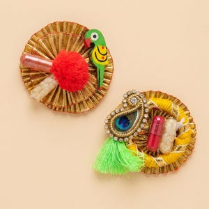 Op de afbeelding: Twee decoratieve, cirkelvormige ornamenten. Eén heeft een rode pompon, een roze cilinder en een houten papegaai. De andere heeft een pauwenveerontwerp, een groene kwast en twee capsulevormige objecten. Beide hebben gouden en gele accenten.