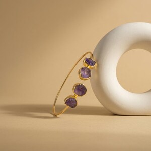 May include: A gold-toned open cuff bracelet featuring four raw, purple amethyst stones set in metal bezels. The bracelet has a thin, circular band design, with two stones positioned at each end of the opening, showcasing natural, irregular shapes.
