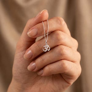 Peut inclure: Un collier de couleur argentée doté d'un petit pendentif délicat en forme de symbole Om. Le pendentif est suspendu à une fine chaîne assortie de couleur argentée, créant une esthétique simple et épurée pour ce bijou.