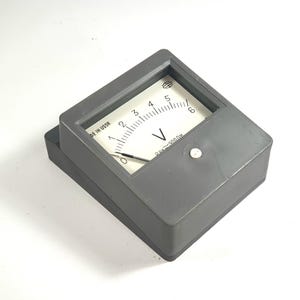 Puede incluir: Un voltímetro analógico gris con una esfera blanca y marcas negras. El medidor mide en voltios, con un rango de 0 a 6. El texto "MADE IN USSR" es visible. Hay un pequeño botón en la parte delantera.