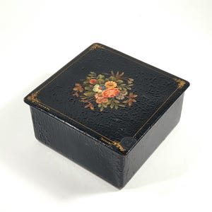 May include: Black square box with a textured surface, adorned with a floral bouquet on the lid. The flowers are in shades of orange, yellow, blue, and pink. Gold trim accents the edges.