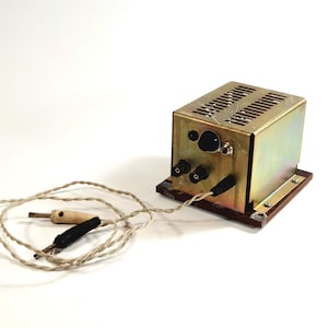 Op de afbeelding: Een vintage goudkleurig elektrisch apparaat met een rechthoekige vorm en een sleufvormige bovenkant. Het staat op een houten voet en heeft verschillende zwarte knoppen en een zwart-witte draad met twee connectoren. Het apparaat is waarschijnlijk uit het midden van de 20e eeuw.
