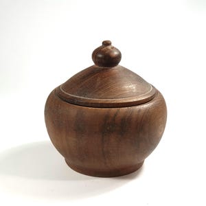 Op de afbeelding: Een handgemaakte houten pot met een deksel. De pot heeft een warme bruine kleur met een gladde, gepolijste afwerking. Het deksel heeft een kleine, afgeronde knop. De pot wordt waarschijnlijk gebruikt voor opslag of decoratie.