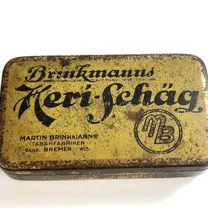 Può includere: Scatola rettangolare vintage con finitura dorata e marrone anticata. La scatola presenta il testo "Brinkmanns Keri-Schäg" in corsivo nero, insieme a testo aggiuntivo e un logo. Probabilmente un contenitore per tabacco o tabacco da fiuto.