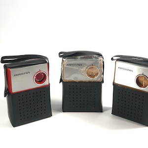 Op de afbeelding: Drie vintage draagbare radio's in zwarte leren hoesjes. De radio's hebben zilveren en rode of gouden voorkanten met een afstemknop en de tekst "SOUNDESIGN". De hoesjes hebben een handvat en een geperforeerde voorkant.