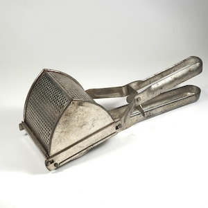 May include: A vintage potato ricer made of silver-colored metal. The ricer has a perforated, semi-circular basket and long handles for pressing. This kitchen utensil is designed for mashing potatoes and other soft foods.