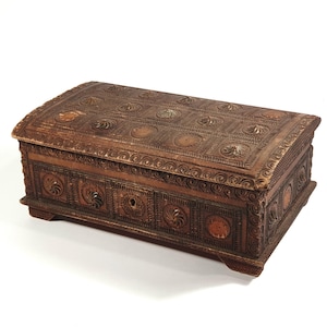 May include: A rectangular wooden chest with a domed lid. The chest is intricately carved with floral and geometric patterns. The wood has a warm, brown tone, and the chest appears to be antique or vintage. It has a keyhole and a small knob.