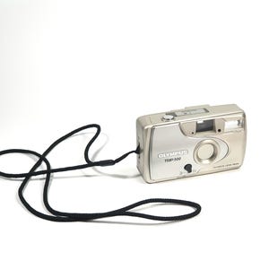 Könnte beinhalten: Silberne Olympus Trip 500 Filmkamera mit schwarzem Halsband. Die Kamera hat ein Objektiv, einen Blitz und verschiedene Knöpfe. Die Kamera befindet sich auf weißem Hintergrund.