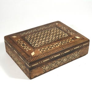 May include: A rectangular wooden box with detailed inlaid patterns. The lid has a central geometric design, surrounded by floral and geometric borders. The box is mainly brown with lighter wood accents.