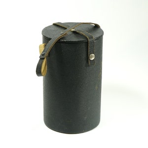 Puede incluir: Una caja cilíndrica de cuero negro con una correa de cuero marrón y cierres metálicos. La caja tiene un asa y está cerrada.