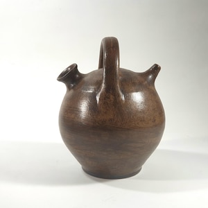 May include: A brown ceramic jug with a rounded body, two spouts, and a handle. The jug has a smooth, polished surface and a dark brown color. It is likely used for holding liquids.