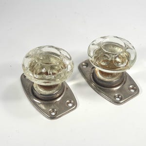 Puede incluir: Dos pomos de puerta de vidrio vintage con un diseño claro y texturizado. Cada pomo está montado sobre una placa trasera rectangular de metal plateado con cuatro orificios para tornillos. Los pomos son probablemente para puertas interiores.