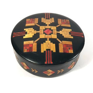 May include: A round, black wooden box with a geometric design inlaid in red, gold, and brown wood. The design is a stylized starburst pattern.