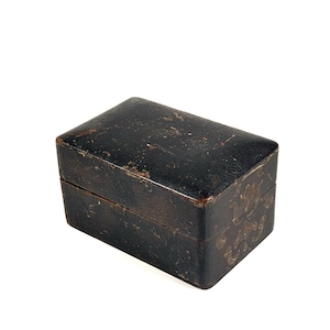 Può includere: Una scatola nera rettangolare antica con coperchio incernierato. La scatola ha un aspetto usurato e strutturato con segni visibili di età e utilizzo. La scatola è probabilmente realizzata in pelle o materiale simile.