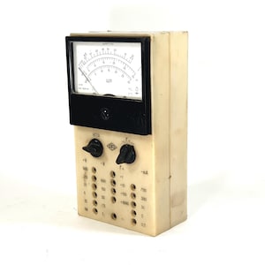 Puede incluir: Un multímetro analógico vintage con una cara blanca y un dial negro. El medidor tiene una perilla selectora de rango y una perilla selectora de función. El medidor tiene una cara en blanco y negro con el texto "IC10" y "V~" impreso en él. El medidor tiene un rango de 0,3 a 1000 mA y de 0,3 a 1000 V.