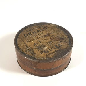 Op de afbeelding: Een ronde metalen doos in vintage-stijl met een verweerde goud- en bruinkleurige afwerking. Het deksel bevat zwarte tekst met PHARMACIE DEHAUT, No 14-7, Rue du Faubg S. Denis Paris en PILULES purgatives, vergezeld van botanische illustraties.
