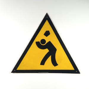 Può includere: Un segnale di avvertimento triangolare giallo con un bordo nero spesso. Il centro presenta una silhouette grafica nera di una persona che solleva una pietra sopra la testa, indicando un pericolo di caduta di detriti.