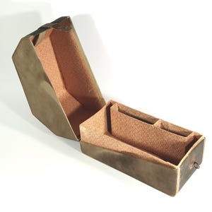 Puede incluir: Una caja abierta de estilo vintage con tapa con bisagras. El exterior es de color verde oliva apagado, mientras que el interior está forrado con una tela estampada de color melocotón. La caja tiene un pequeño compartimento y un anillo de metal en el lateral.