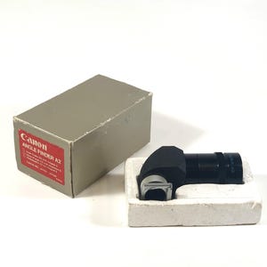 Puede incluir: Un visor de ángulo Canon A2 negro en su caja original. La caja es gris con letras rojas. El visor es un pequeño dispositivo de mano con una empuñadura de goma negra y una lente plateada.