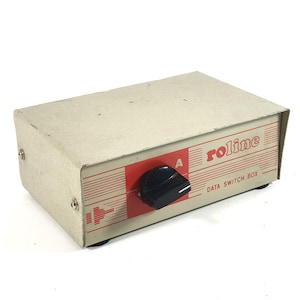 Puede incluir: Una caja de conmutación de datos rectangular de metal beige con un mando giratorio negro central sobre un panel rojo. El panel frontal muestra el texto rojo roline y DATA SWITCH BOX, con una letra A sobre el mando y líneas.