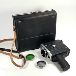 May include: A vintage black and silver Super 8 film camera with a black leather case and a brown leather strap. Two circular lens filters, one green and one clear, are also included.