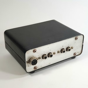 Op de afbeelding: Een zwart rechthoekig audio-apparaat met een zilveren paneel met meerdere ingangs- en uitgangsconnectoren. Het apparaat heeft een afgeronde bovenkant en is gemonteerd op kleine zwarte voetjes. Het paneel heeft de labels "L", "R" en "IN".