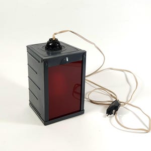 Op de afbeelding: Een grijze metalen donkere kamer veiligheidslamp met een rood filter en een snoer met een stekker.