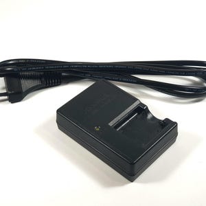 May include: A black battery charger and power cable. The charger is rectangular with a recessed area for a battery. The power cable is black with a two-pin plug.