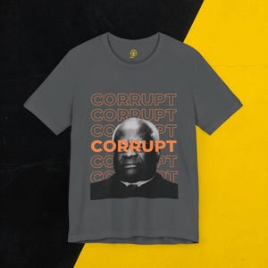 May include: A dark grey t-shirt featuring a black and white image of a man with the word "CORRUPT" repeated in orange lettering.