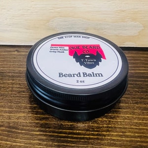 May include: A black tin container with a white label that reads "OKIE BEARD CO." and "Beard Balm". The label also includes the text "One Stop Man Shop", "Ocean Mist, Warm Amber, Crisp Musk", and "T-Town Vibes". The container is 2 oz.