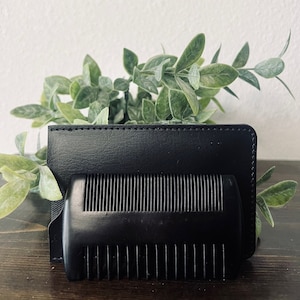 May include: A black wooden beard comb with a rounded handle and fine teeth, paired with a black leather cardholder. The comb and cardholder are set against a backdrop of green foliage.
